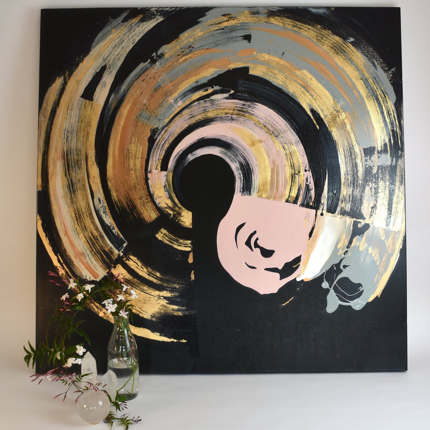 A canvas painting with abstract elements in black, gold, and pink hues, with a smaller pink shape resembling a face or figure in the center. The artwork is accompanied by a small vase with flowers to the bottom left.