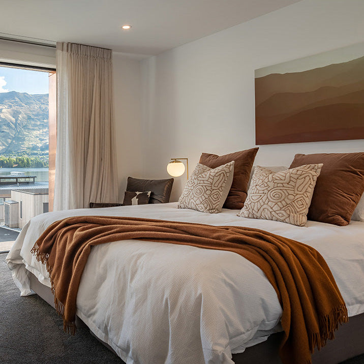 A styled bedroom scene with a bed dressed in white bedding and a rust-brown throw blanket, surrounded by patterned and solid brown pillows. A large window on the left shows a mountain view, and a minimalist landscape painting hangs on the wall above the bed.