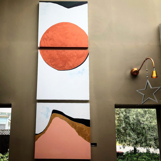 A triptych artwork with a large orange circle on a white background above a canvas with a mountain-like form in beige and brown tones, displayed on a wall.